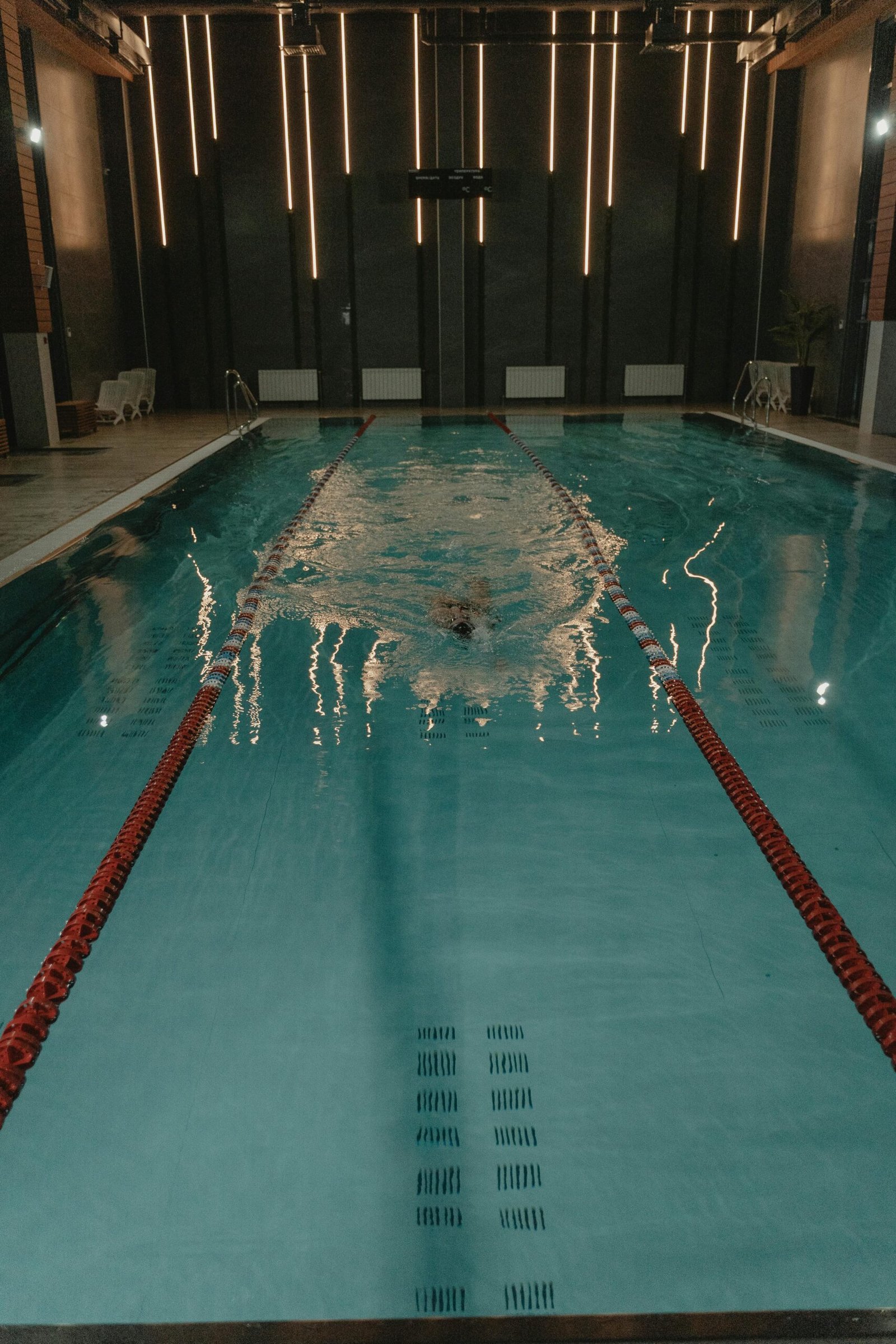 An unrecognizable swimmer glides through an indoor pool with clear pool dividers and glowing lights.